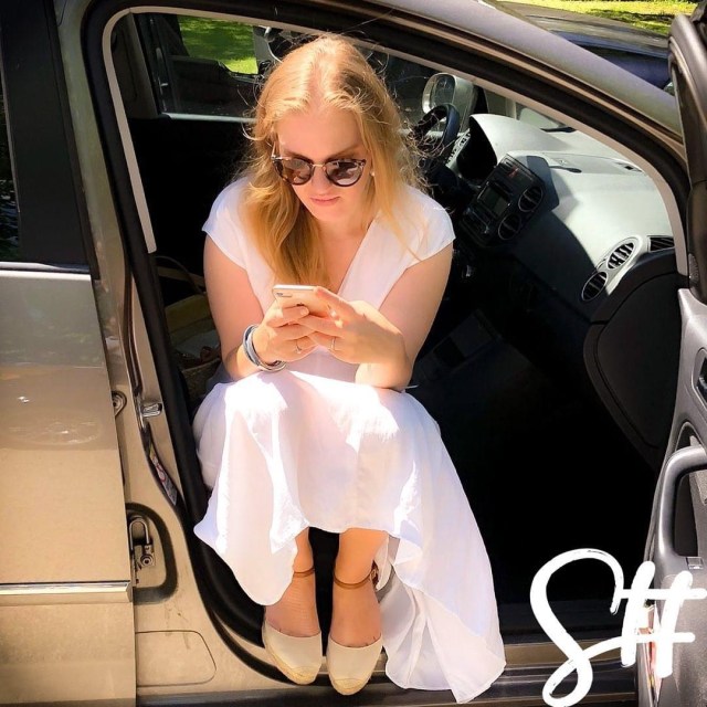 Blonde Frau in weiß sitzt Autotür und tippt in das Handy